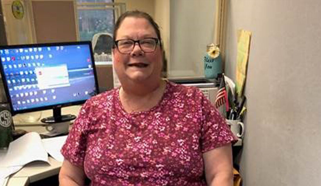 Carol, smiling, with her prosthetic at her computer desk.